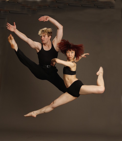 One male dancer in a jump with his legs splayed. His female counterpart leaps in the air with her front leg straight and back leg in attitude. She looks over her shoulder